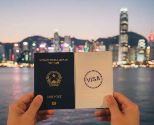 Một người cầm hộ chiếu có visa Hồng Kông trước khung cảnh vịnh Victoria và các tòa nhà chọc trời.