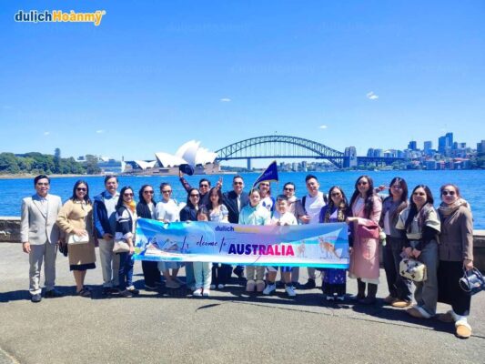 Đoàn khách của Du Lịch Hoàn Mỹ chụp ảnh kỷ niệm tại Sydney, Úc, với hậu cảnh là Nhà hát Opera và Cầu cảng Sydney, thể hiện một chuyến đi thành công và vui vẻ.
