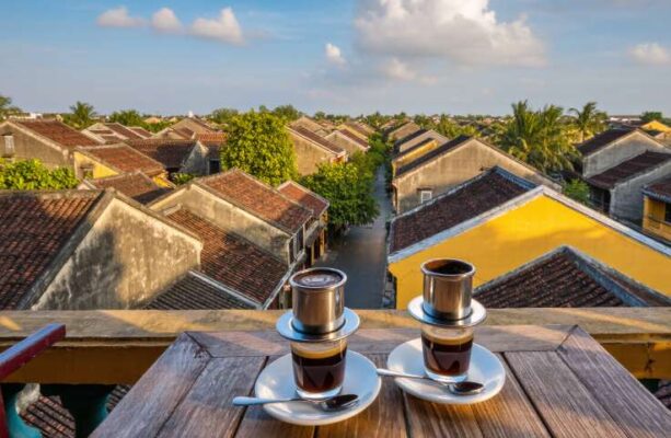 Hai tách cà phê trên bàn gỗ tại một quán cà phê sân thượng, nhìn xuống những mái ngói rêu phong của Phố cổ Hội An.